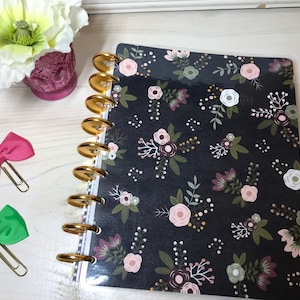 May include: A black planner with a floral pattern featuring pink, white, and green flowers. The planner has gold rings and a clear plastic cover.