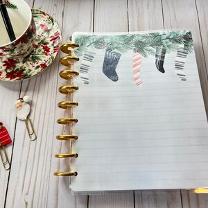 May include: A white planner with a gold coil binding and a Christmas stocking design on the cover. The cover features a green pine branch with red and white striped stockings hanging from it. The planner has lined pages and the text "Live & Remember" is printed on the bottom of the cover.