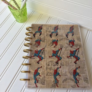 May include: A spiral-bound notebook featuring a repeating pattern of Spider-Man in action poses. The cover has a clear overlay and gold-colored spiral binding. The background is a comic book style design with various panels.
