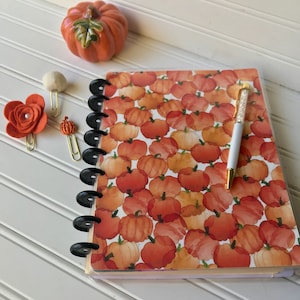 May include: A black and white spiral bound notebook with a pumpkin pattern. The cover features a watercolor design of orange pumpkins on a white background.