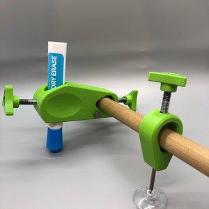 May include: A green 3D printed clamp with a suction cup base holds a wooden dowel. A dry erase marker is held in place by another green clamp attached to the dowel.