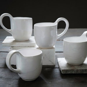May include: Four white ceramic mugs with distinctive, curved handles. The mugs are presented on square, light-coloured stone blocks and a dark wooden surface. The mugs have a simple, modern design, suitable for coffee or tea.
