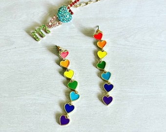 May include: A pair of rainbow heart dangle earrings with a gold chain and a rainbow heart pendant necklace. The hearts are red, orange, yellow, green, blue, indigo, and violet.