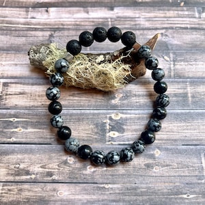 May include: A black and white speckled stone bracelet with black lava beads and silver metal accents. The bracelet is on a wooden surface.