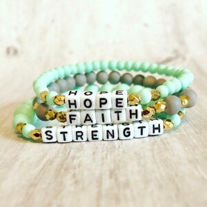 May include: A set of three bracelets with beads in mint green and gray colors. The bracelets have gold accents and white letter beads spelling out the words "HOPE", "FAITH", and "STRENGTH".