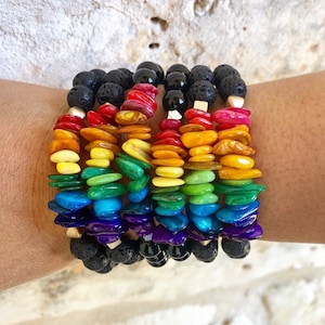 May include: A set of seven rainbow-colored bracelets made of small, flat, shell-like beads. The bracelets are stacked on a person's wrist, with the top bracelet being red, followed by orange, yellow, green, blue, purple, and black. The black bracelet is made of lava rock beads.