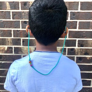 Op de afbeelding: Een jong persoon draagt een wit T-shirt met een turquoise kralenketting waarop "MOM" in kleurrijke letters staat. De ketting is bevestigd aan een koptelefoon.