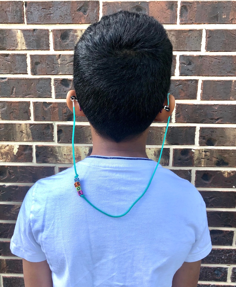 Op de afbeelding: Een jong persoon draagt een wit T-shirt met een turquoise kralenketting waarop "MOM" in kleurrijke letters staat. De ketting is bevestigd aan een koptelefoon.