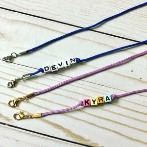 Op de afbeelding: Drie kleurrijke kralenkettingen met metalen sluitingen. De kettingen spellen de namen "Devin" en "Kyra" uit.