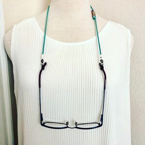 Op de afbeelding: Een turquoise blauwe brilketting met gekleurde kralen en zilveren clips. De ketting houdt een paar zwarte gemonteerde brillen vast.