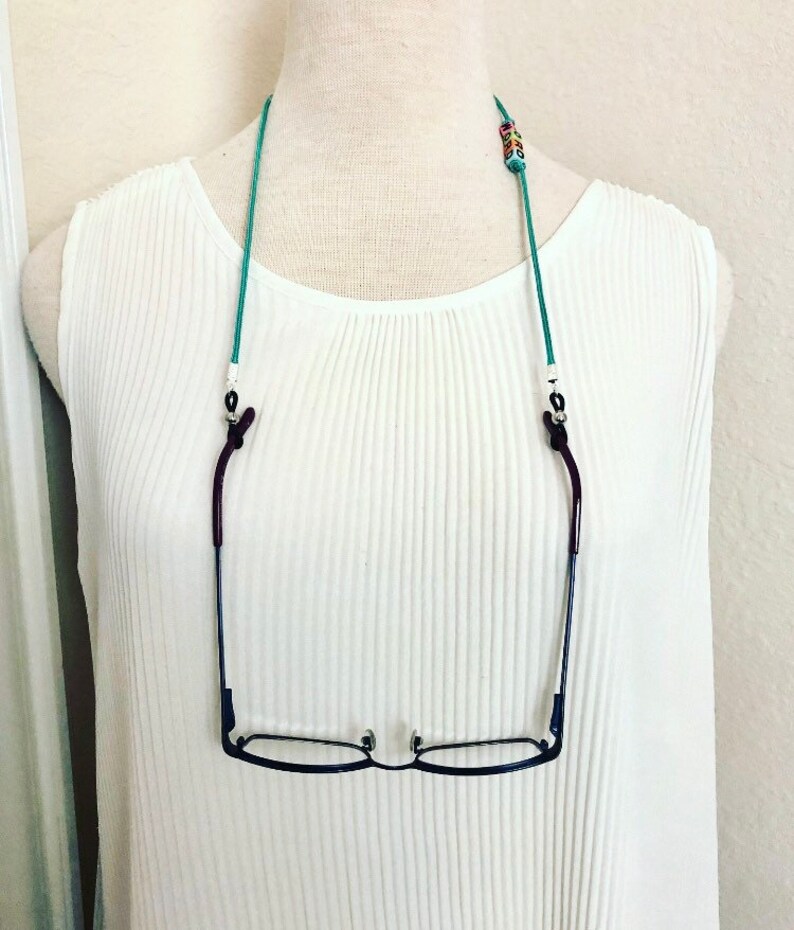Op de afbeelding: Een turquoise blauwe brilketting met gekleurde kralen en zilveren clips. De ketting houdt een paar zwarte gemonteerde brillen vast.