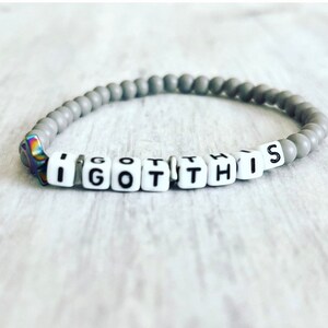 May include: A gray beaded bracelet with white letter beads spelling out "I GOT THIS".