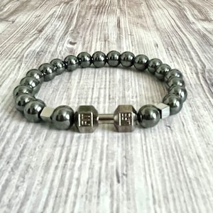 May include: A black hematite beaded bracelet with a silver dumbbell charm. The bracelet is on a light wood background.