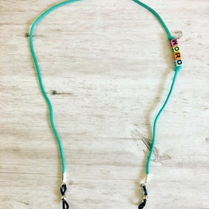 Op de afbeelding: Een turquoise blauwe brilketting met zwarte koorduiteinden en zilveren sluitingen. De ketting heeft kleurrijke letterkraaltjes die het woord "WORD" spellen.