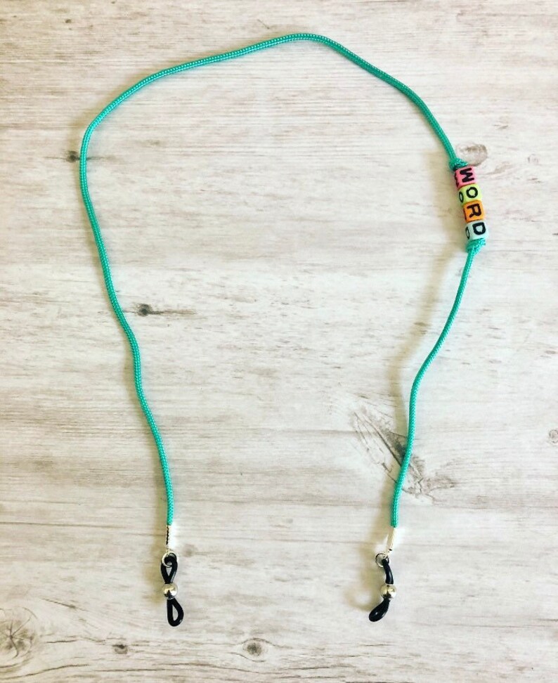 Op de afbeelding: Een turquoise blauwe brilketting met zwarte koorduiteinden en zilveren sluitingen. De ketting heeft kleurrijke letterkraaltjes die het woord "WORD" spellen.
