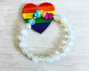 May include: A white opal beaded bracelet with a rainbow heart charm. The charm has a rainbow stripe design and a row of colorful beads.