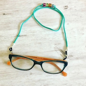 Op de afbeelding: Turquoise blauwe brilketting met zwart en zilveren beslag. De ketting heeft gekleurde kralen die het woord "WORD" spellen. De ketting is bevestigd aan een bruin en zwart bril.