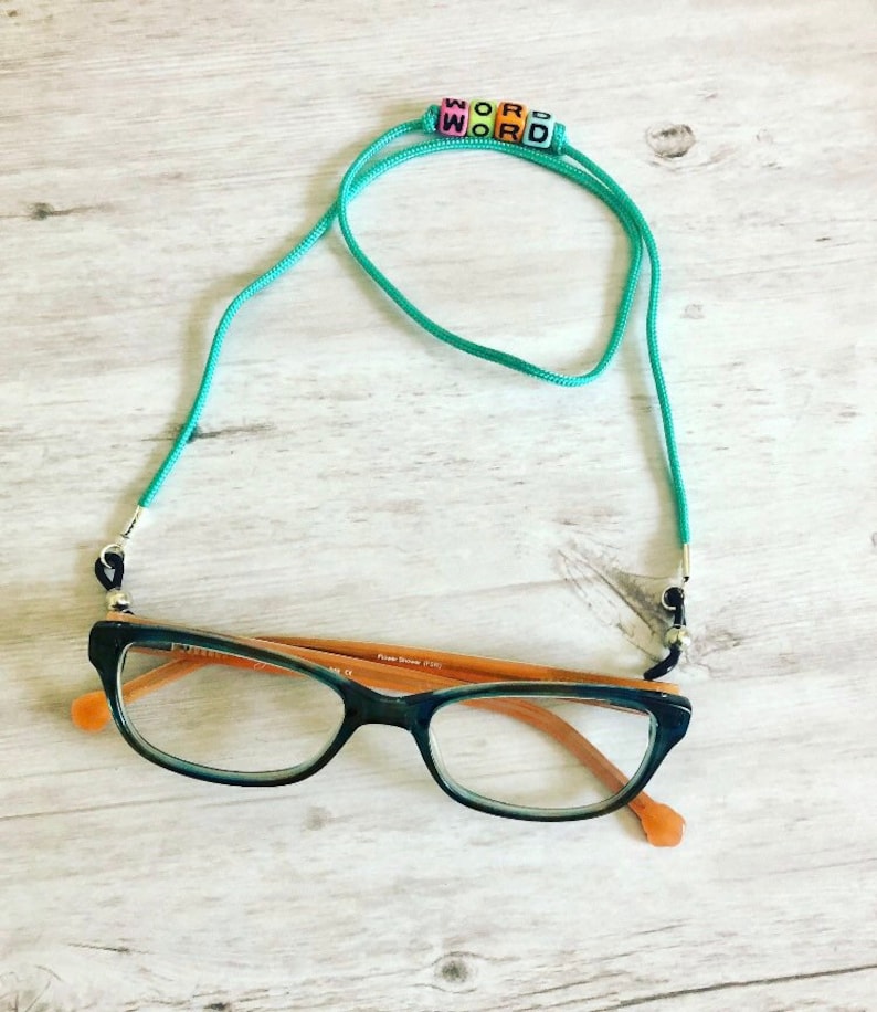 Op de afbeelding: Turquoise blauwe brilketting met zwart en zilveren beslag. De ketting heeft gekleurde kralen die het woord "WORD" spellen. De ketting is bevestigd aan een bruin en zwart bril.