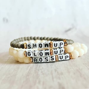 May include: Three stacked bracelets with white beads and gold accents. The beads have the words "SHOW UP", "GLOW UP", and "BOSS UP" spelled out.