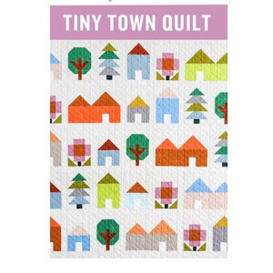 May include: A quilted fabric featuring a repeating pattern of colorful houses, trees, and flowers. The quilt has a white background with a grid pattern. The top of the quilt has a lavender banner that reads "TINY TOWN QUILT".