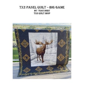 May include: A quilt featuring a brown moose in a snowy forest scene. The quilt is framed with a dark blue background and a gold and green diamond pattern. The quilt is titled "TX2 Panel Quilt - Big Game" and "By: Traci Drew TX2 Quilt Shop".