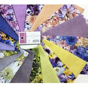 May include: A collection of fabric squares arranged in a circular pattern. The fabrics feature floral patterns in shades of blue, purple, yellow, and peach. A small card with text is in the center. The fabrics are likely for quilting or crafting.