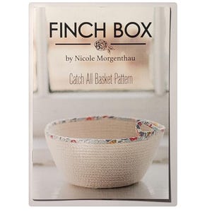 May include: A white fabric basket with a floral print trim. The basket is labeled "Finch Box by Nicole Morgenthau Catch All Basket Pattern".