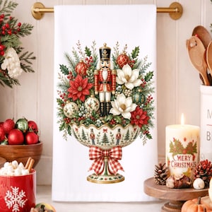 May include: A white kitchen towel featuring a central illustration of a decorative pedestal bowl filled with a nutcracker, poinsettias, white magnolia flowers, pine branches, and ornaments, accented with a red and white checkered bow.