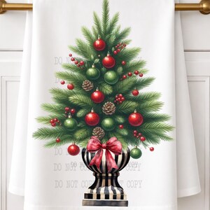 May include: A white kitchen towel featuring a central illustration of a green pine Christmas tree decorated with red and green ornaments, pinecones, and red berries. The tree sits in a black and white striped urn adorned with a red bow.