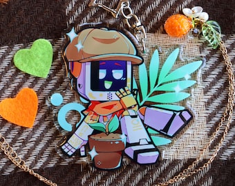 Puede incluir: Un amuleto colorido con un robot de dibujos animados que lleva un sombrero marrón y sostiene una planta en maceta. El robot tiene un cuerpo blanco y morado con una cara negra y manos amarillas. El amuleto está unido a una cadena dorada.