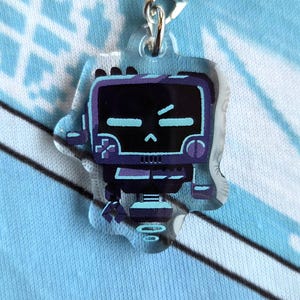 May include: A clear acrylic charm with a robot design in purple and teal. The robot has a screen-like face. It has a silver clasp and a black cord. The background is a light blue fabric. The charm is designed to be attached to a bag or phone.