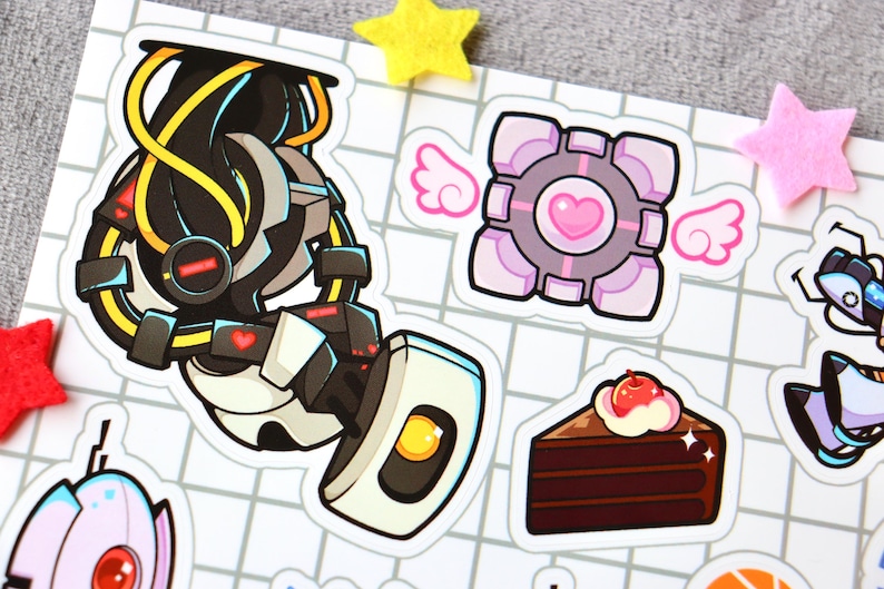 Puede incluir: Un conjunto de pegatinas con personajes del videojuego Portal. Las pegatinas son coloridas y presentan un robot, un cubo, un pastel y un coraz&oacute;n.