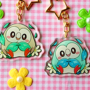 Puede incluir: Dos llaveros de acrílico transparente con ilustraciones de dibujos animados de un personaje de Pokémon parecido a un búho, uno marrón y otro azul. Ambos personajes sostienen hojas verdes en sus manos y tienen una margarita blanca a cada lado.