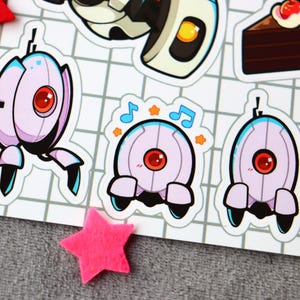 Puede incluir: Un conjunto de cinco pegatinas con personajes del videojuego Portal. Las pegatinas est&aacute;n impresas en papel blanco y presentan un robot con un ojo rojo, un robot con un ojo azul, un robot con un ojo rojo y notas musicales, un robot con un ojo azul y un pastel con una cereza en la parte superior.