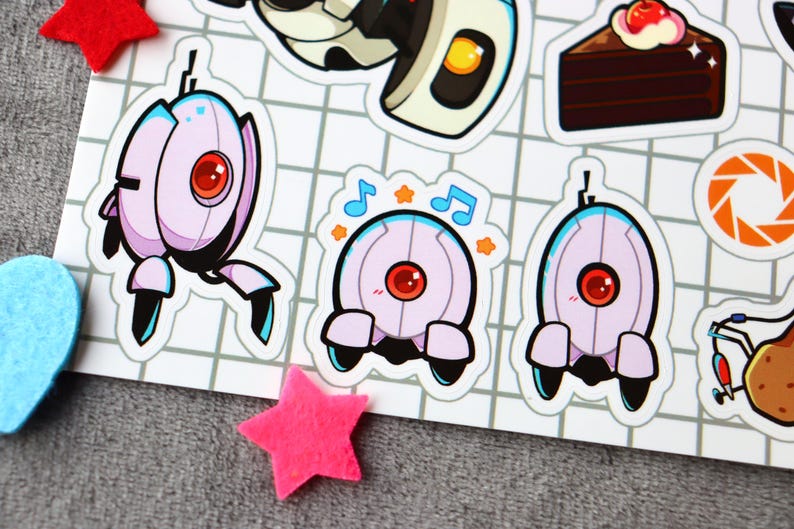 Puede incluir: Un conjunto de cinco pegatinas con personajes del videojuego Portal. Las pegatinas est&aacute;n impresas en papel blanco y presentan un robot con un ojo rojo, un robot con un ojo azul, un robot con un ojo rojo y notas musicales, un robot con un ojo azul y un pastel con una cereza en la parte superior.