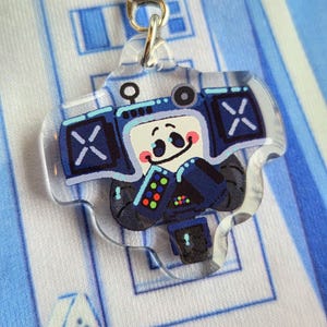 May include: A clear acrylic charm depicting a cartoon character designed as a blue boombox with a smiling face. The charm has a silver-coloured metal clasp for attachment. The background is a light blue and white pattern.