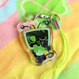 May include: A clear acrylic charm depicting a cartoon robot with a green screen face, a green bow tie, and a microphone. It has a silver clasp and a neon green lanyard. The background is a colourful, rainbow-striped fabric.