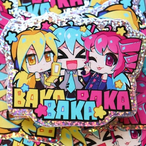 May include: A holographic sticker featuring three chibi-style anime characters with yellow, teal, and pink hair. The characters are positioned above the repeated text BAKA in bold yellow, pink, and blue block letters, accented by small stars.