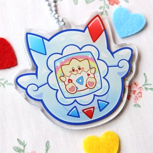 Puede incluir: Un llavero de acrílico azul y blanco con un personaje de dibujos animados con un cuerpo amarillo y rosa dentro de un marco azul y blanco. El personaje sostiene un objeto azul y blanco con un corazón rosa. El llavero tiene una oreja roja y azul en el lado izquierdo y una oreja roja en el lado derecho.