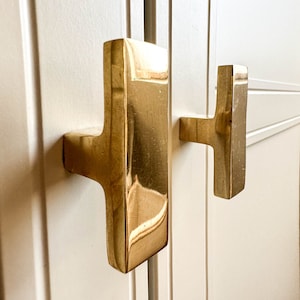 May include: Two gold cabinet door pulls, one is a rectangular shape with a T-shaped handle, the other is a smaller T-shaped handle.