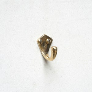 Pode incluir: Um gancho de metal dourado com uma base em forma de diamante e um único gancho. O gancho está montado em uma parede branca.