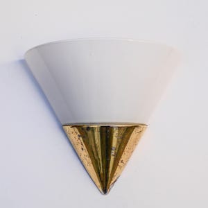 May include: A vintage wall sconce featuring a white, cone-shaped shade and a gold-coloured, triangular base. The sconce is mounted on a white wall. The base has a textured, aged appearance.