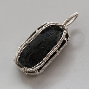 May include: A black tourmaline pendant wrapped in silver wire. The rectangular stone is encased in a woven wire frame with a loop at the top for a chain. The pendant is displayed on a light gray surface.