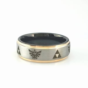 Puede incluir: Un anillo negro y plateado con una banda dorada y un diseño de Trifuerza. El anillo está hecho de carburo de tungsteno.