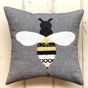 May include: A gray pillow with a black and yellow bee applique. The bee has white wings and a black and yellow striped body.