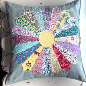 May include: A square throw pillow with a light blue corduroy base featuring a central yellow circular patch surrounded by a sunburst pattern of multi-colored fabric petals. Each petal displays a unique floral, geometric, or polka dot print.