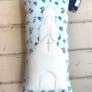 May include: A rectangular pillow with a light blue floral pattern and a white church applique. The church has a cross and a simple design. The pillow is a decorative item.