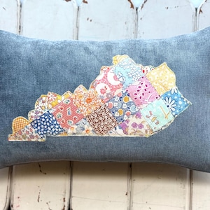 May include: A rectangular denim blue pillow with a patchwork design in the shape of the state of Kentucky. The patchwork features various floral and patterned fabrics in shades of pink, yellow, blue, and brown.