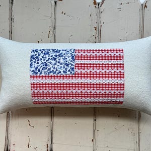 May include: Rectangular cream-colored pillow with a patchwork American flag design. The flag features a blue floral print for the stars and red and white gingham and lace stripes. A patriotic home decor item.