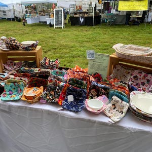 May include: A display of handmade fabric bowls and plates on a white tablecloth at an outdoor market. The bowls feature various colorful patterns, including floral and geometric designs. Some bowls are stacked on wooden crates.